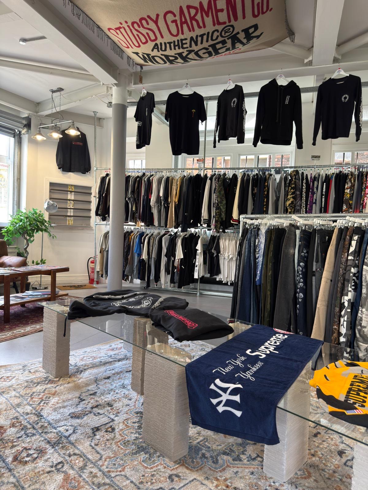 Rails of archive streetwear beneath a St&uuml;ssy banner, with Chrome Hearts pieces hung above a glass plinth.
