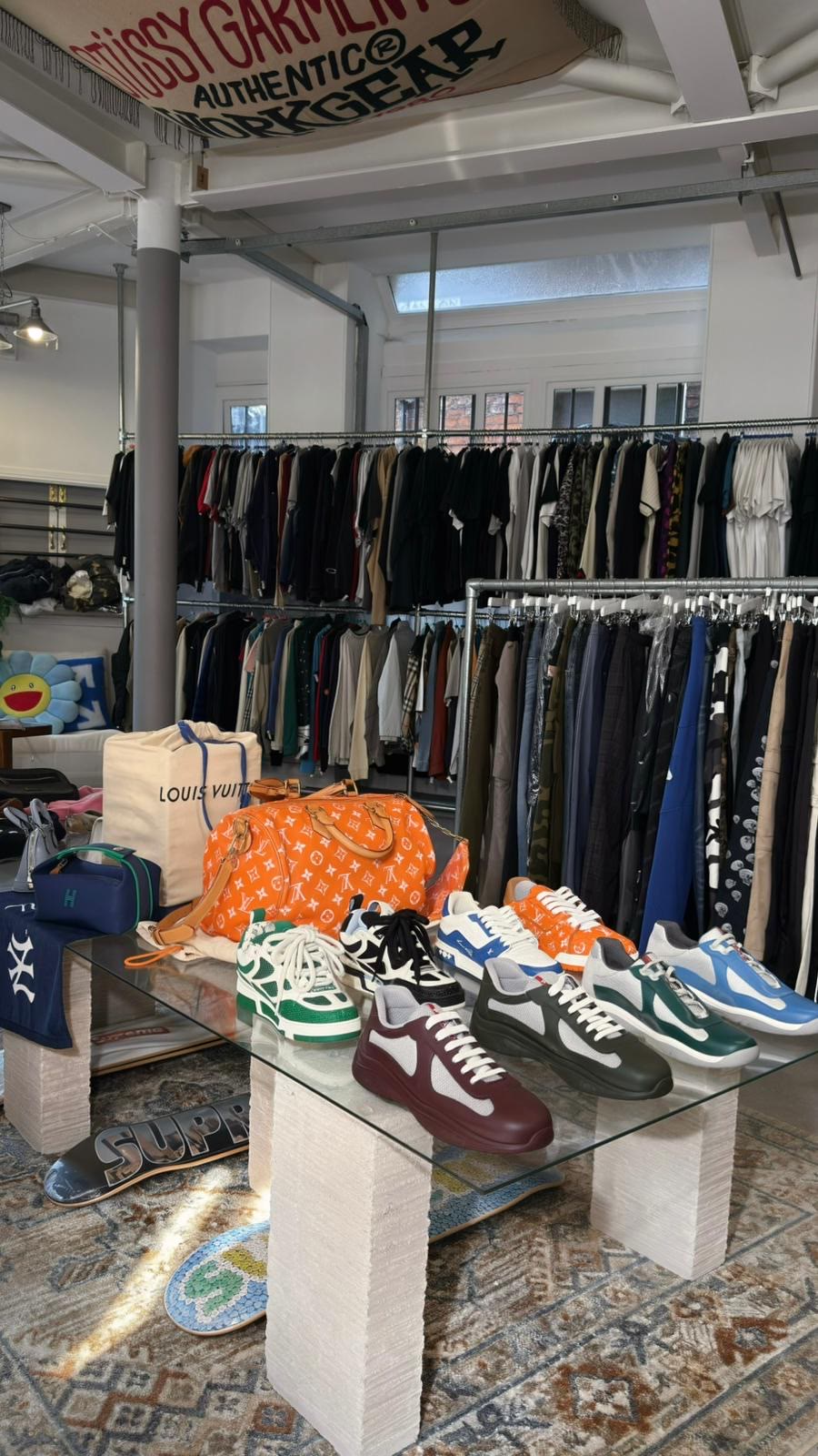 A glass plinth holding Prada America's Cup trainers and a Louis Vuitton holdall, with rails behind.